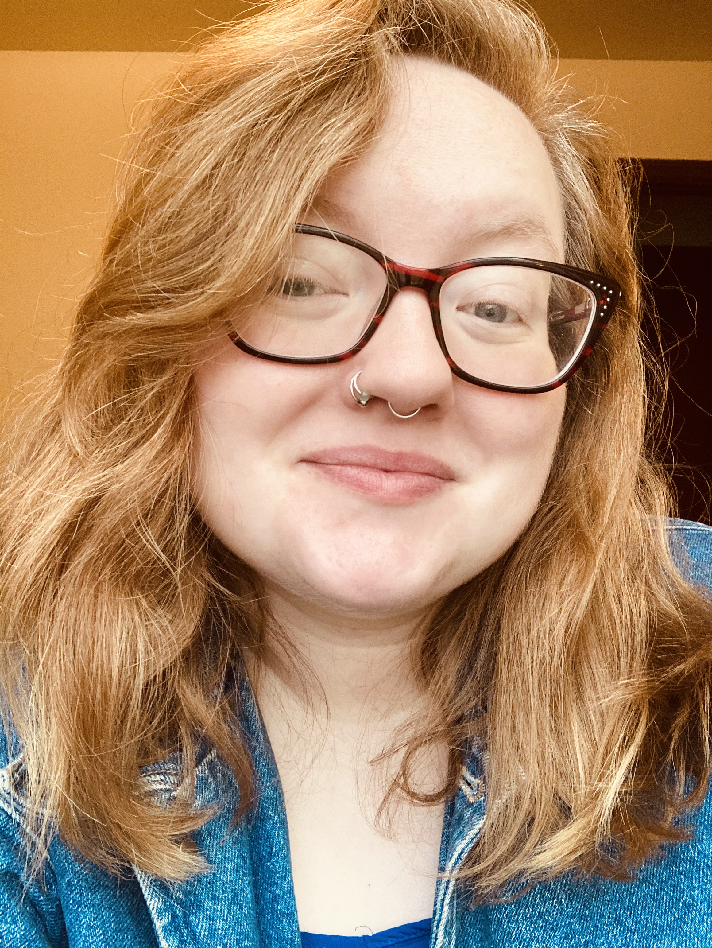 [Image Description: Renn, Director of Operations, a White non-binary person person, smiling at the camera. They have blonde hair, a blue shirt, glasses, and nose piercings.